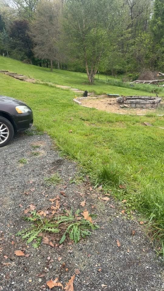 Black car parked on gravel driveway with open field, fire pit, and wooded area in background