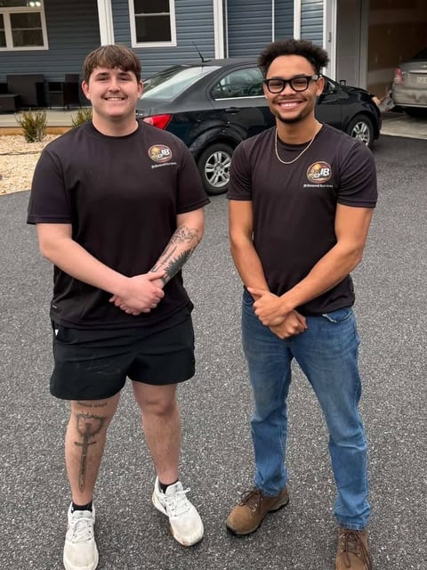 Two men in matching black work shirts stand smiling in a driveway with cars and a house in the background